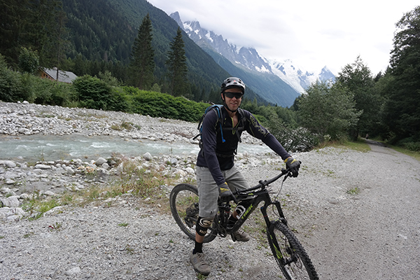 Mountain biker stopped beside river with mountains in the background, smiling at the camera